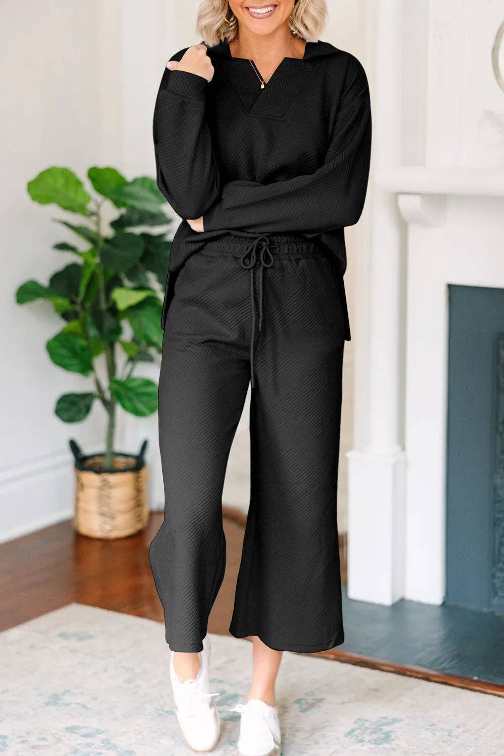 Person wearing a black outfit standing in a room with a plant and fireplace.