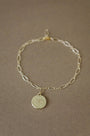 A delicate gold chain bracelet with an oval link pattern and a small round pendant engraved with the number 18, displayed on a textured brown fabric background.