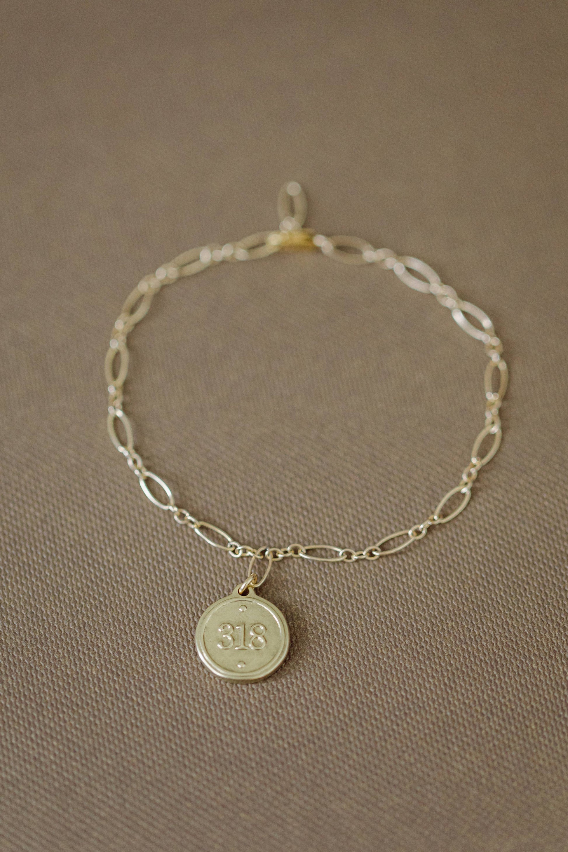A delicate gold chain bracelet with an oval link pattern and a small round pendant engraved with the number 18, displayed on a textured brown fabric background.