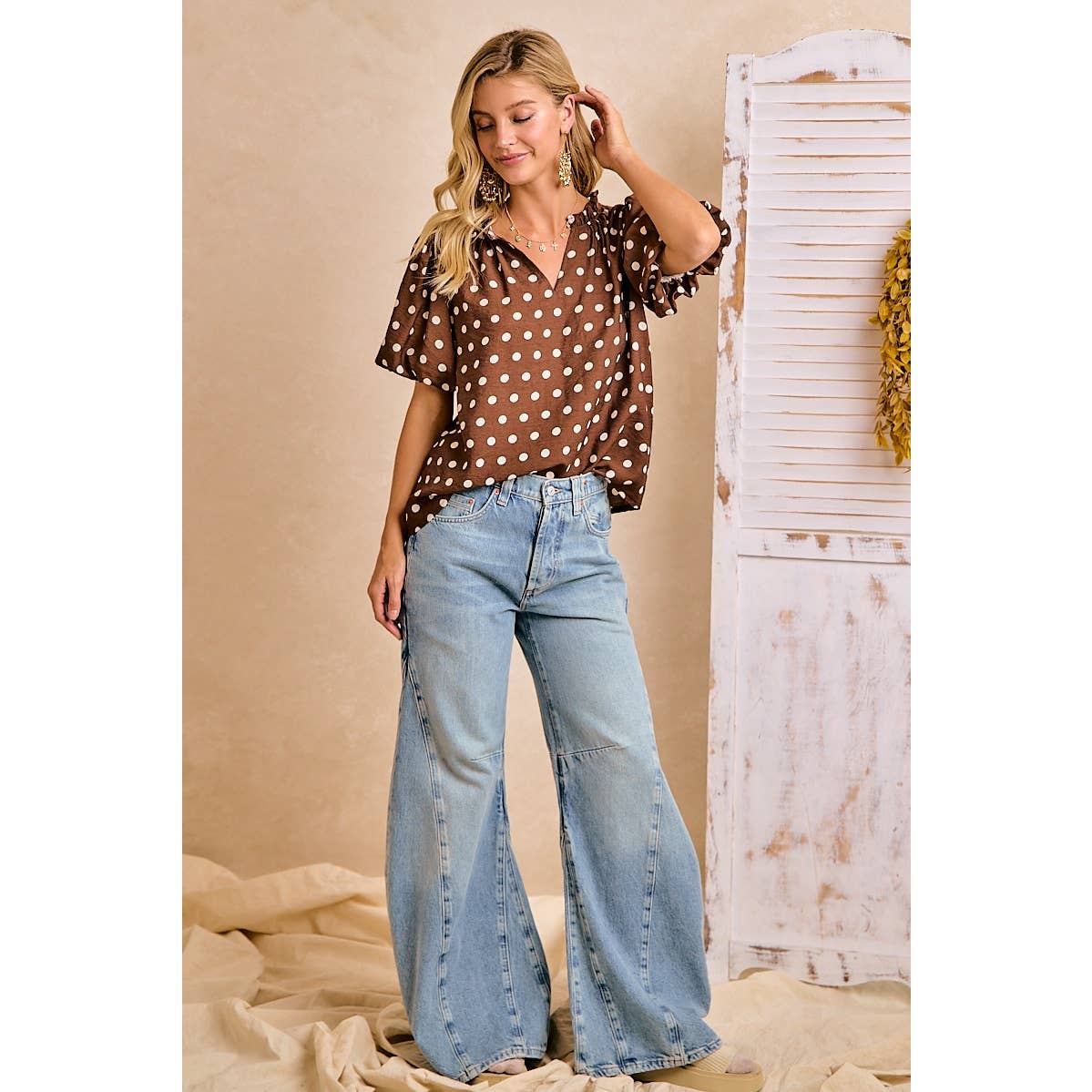 Woman wearing a brown polka dot blouse and light blue jeans against a beige wall.