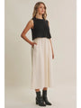 A woman modeling a sleeveless black ribbed knit top paired with a high-waisted, wide-leg cream midi skirt and black ankle boots, set against a plain beige studio background.
