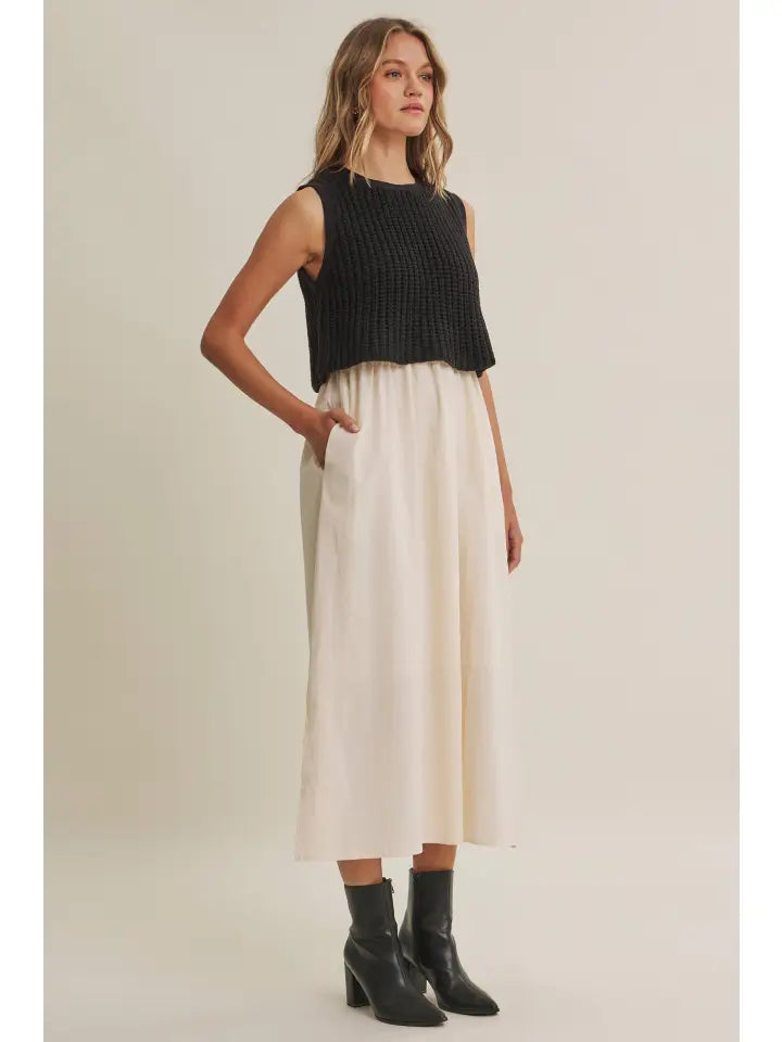 A woman modeling a sleeveless black ribbed knit top paired with a high-waisted, wide-leg cream midi skirt and black ankle boots, set against a plain beige studio background.