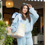 Woman in a light blue outfit holding a striped bag and a drink, standing outdoors.