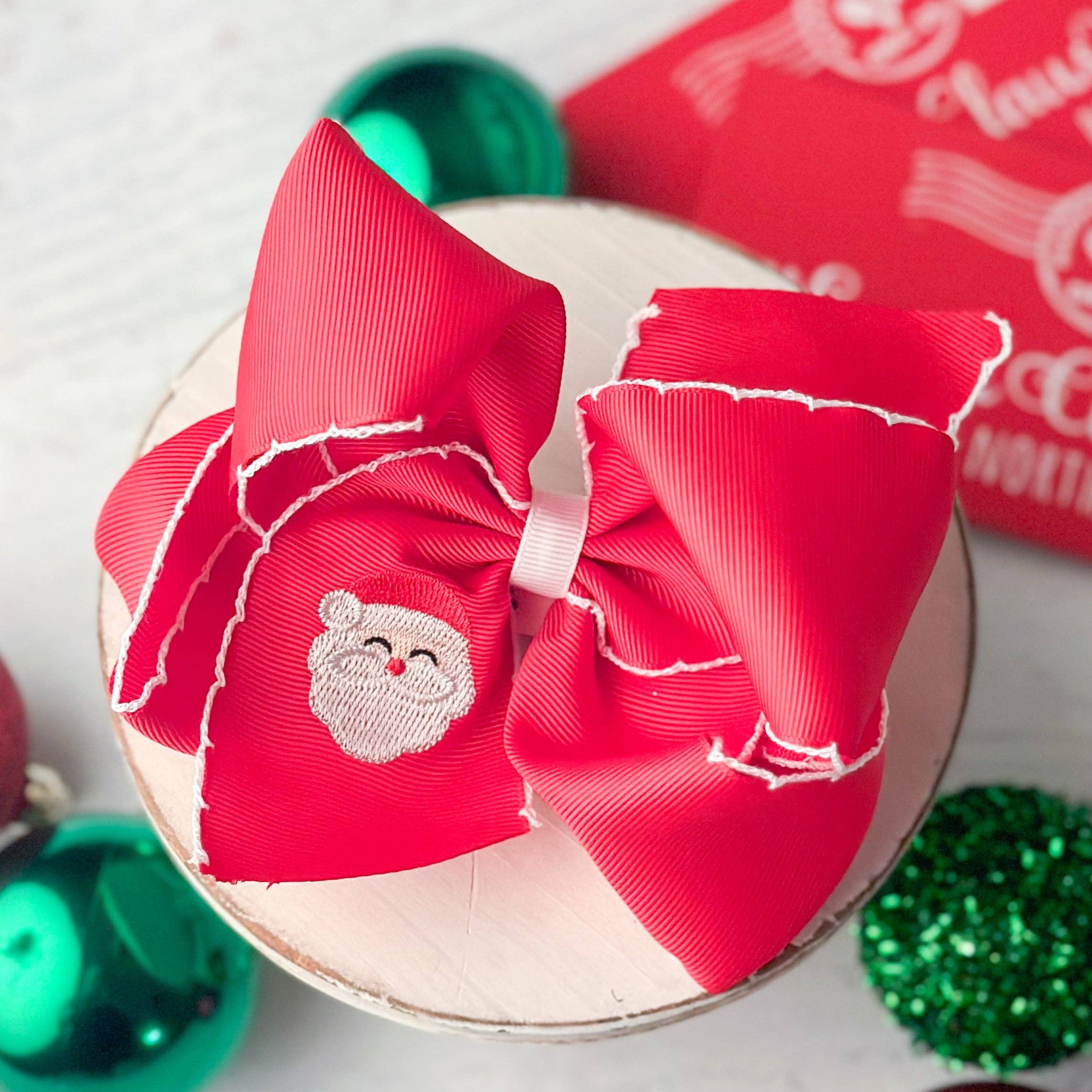 Red hair bow with a Santa Claus patch on a white surface with Christmas decorations.