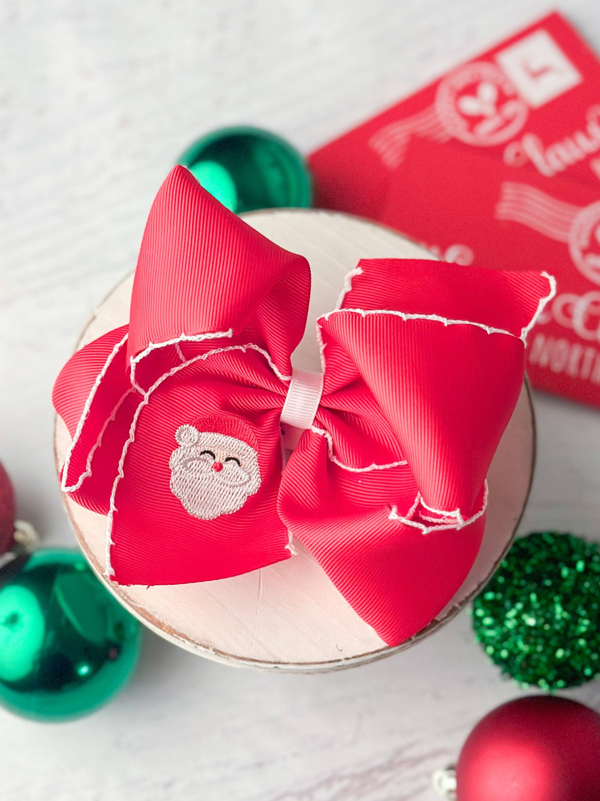 Red hair bow with a Santa Claus patch on a white surface with Christmas decorations.