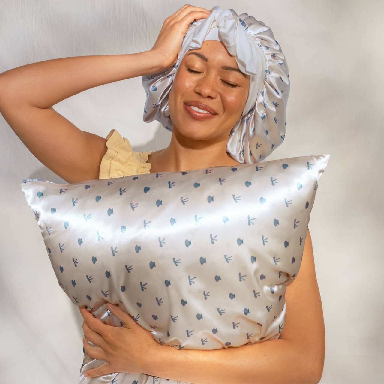Woman wearing a satin headscarf and holding a matching pillow with a floral pattern.