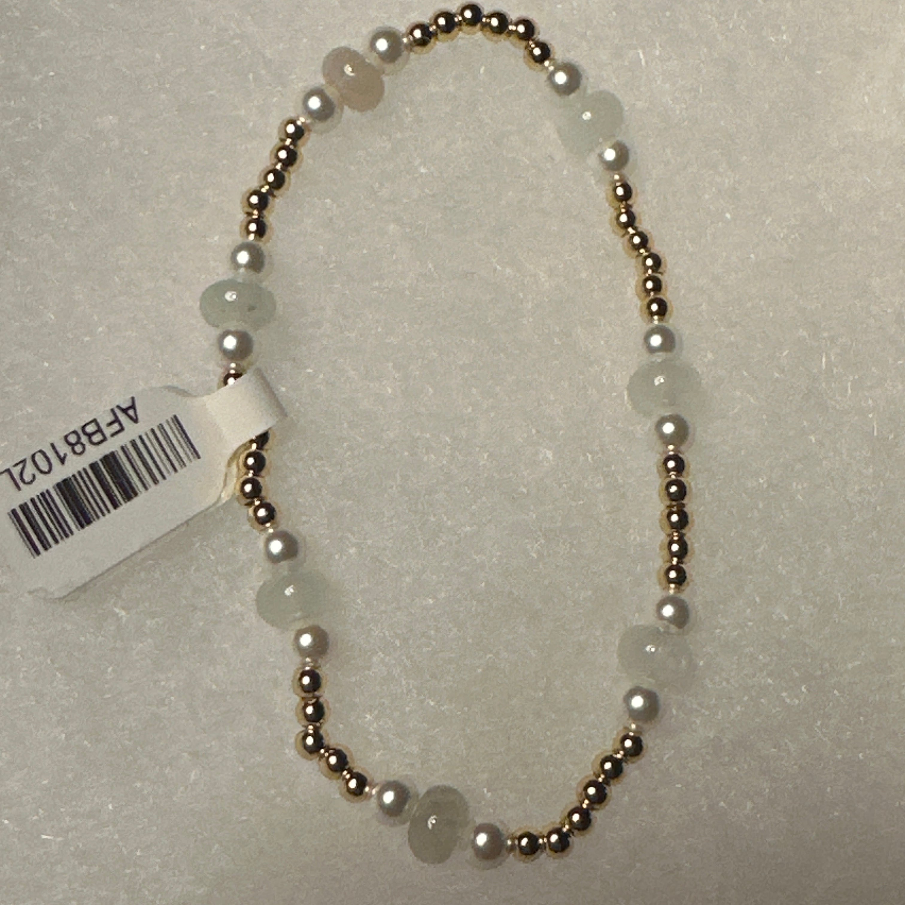 A beaded necklace with round white pearls and pale green stone beads separated by small metallic gold spacer beads, shown on a textured light gray background with a white product tag attached.