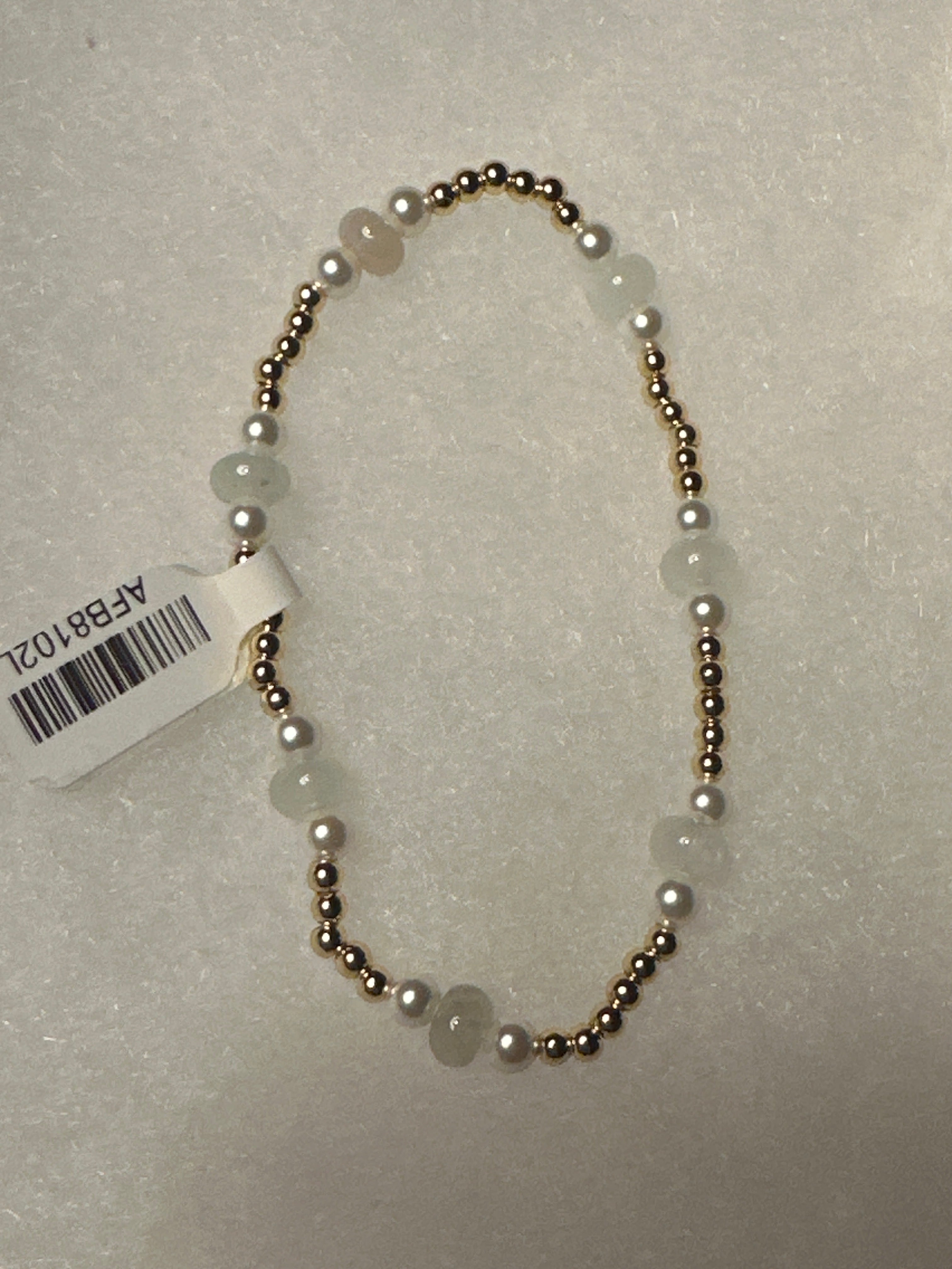 A beaded necklace with round white pearls and pale green stone beads separated by small metallic gold spacer beads, shown on a textured light gray background with a white product tag attached.