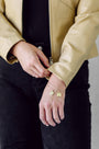 A close-up of a person wearing a gold chain bracelet with a round letter charm and a small square charm, styled with a black top and a beige leather jacket.