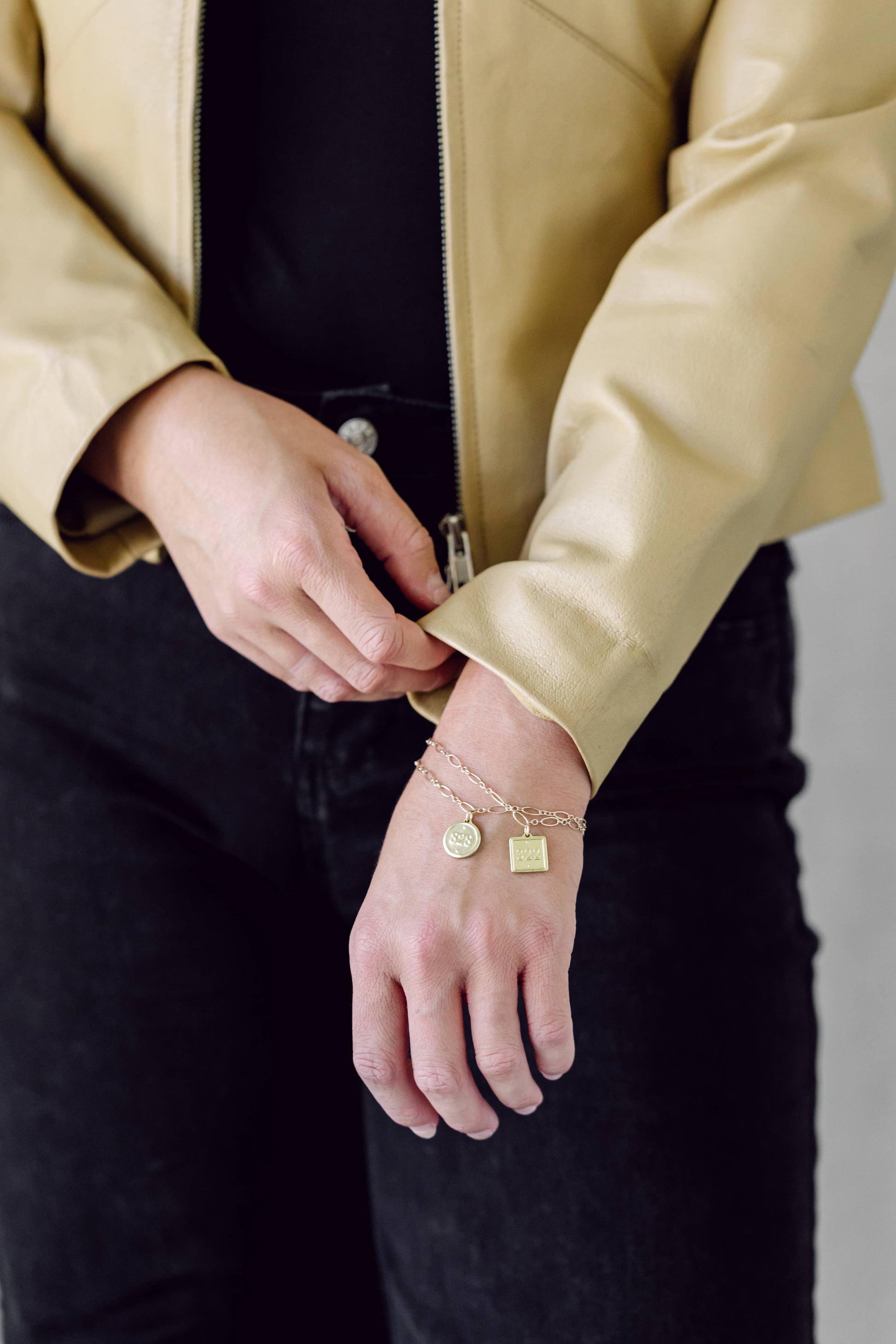 A close-up of a person wearing a gold chain bracelet with a round letter charm and a small square charm, styled with a black top and a beige leather jacket.