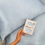 Hand holding a satin pillowcase packaging in front of light blue satin pillows.