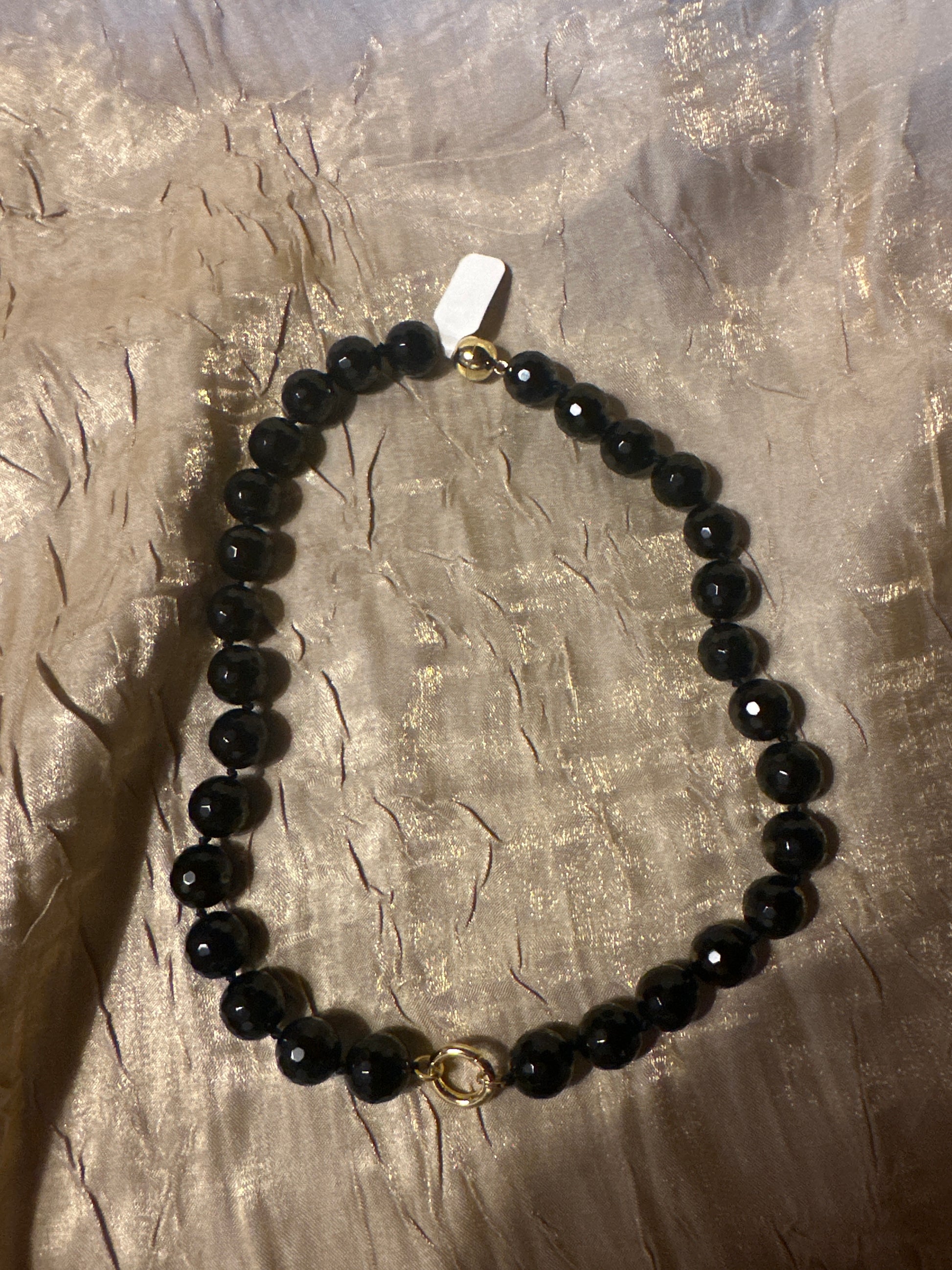 A black beaded necklace with faceted round beads and gold-toned hardware, including a small ring clasp and a white tag, laid on a textured metallic gold surface.