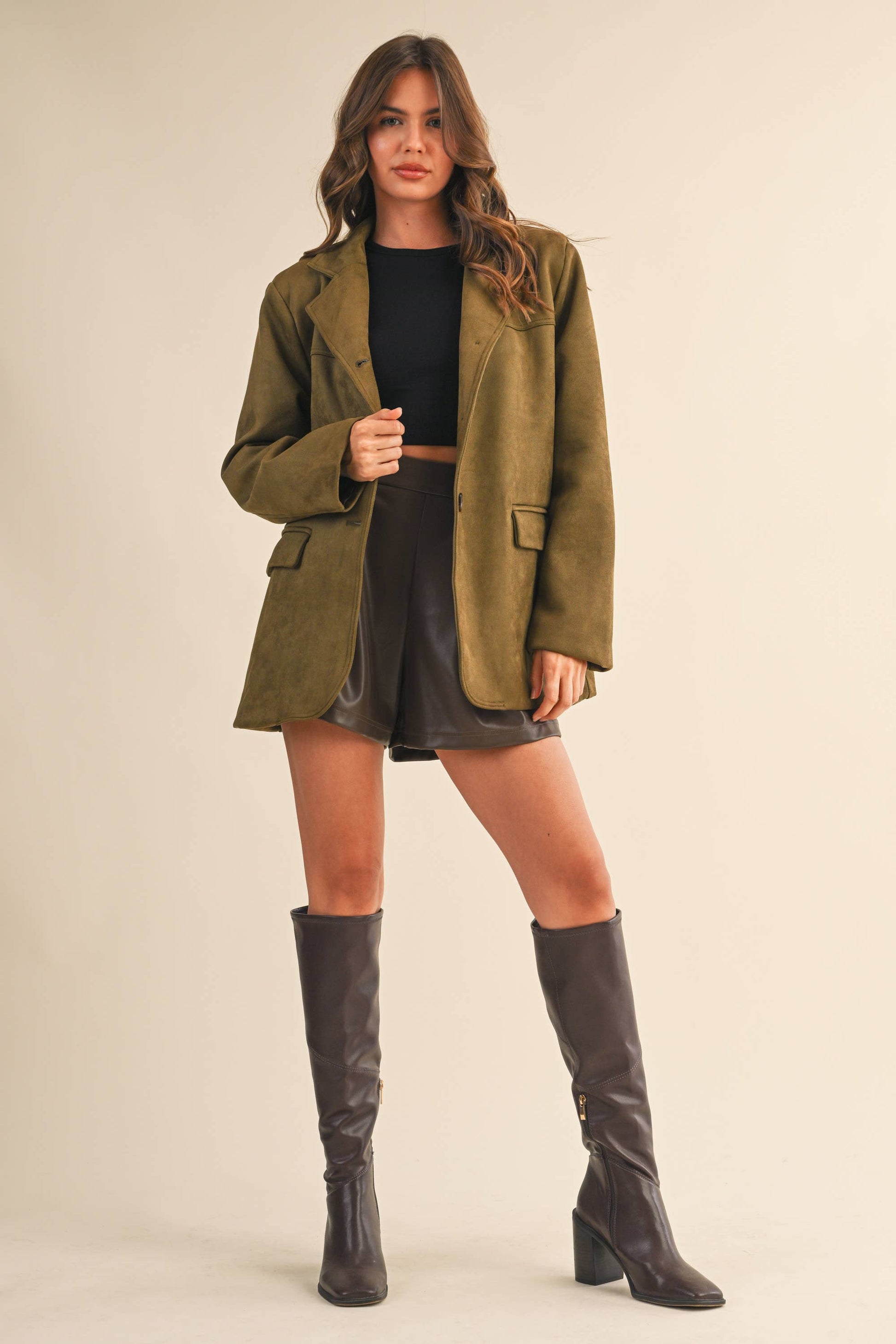 Woman wearing an olive green coat, black top, and knee-high brown boots on a beige background