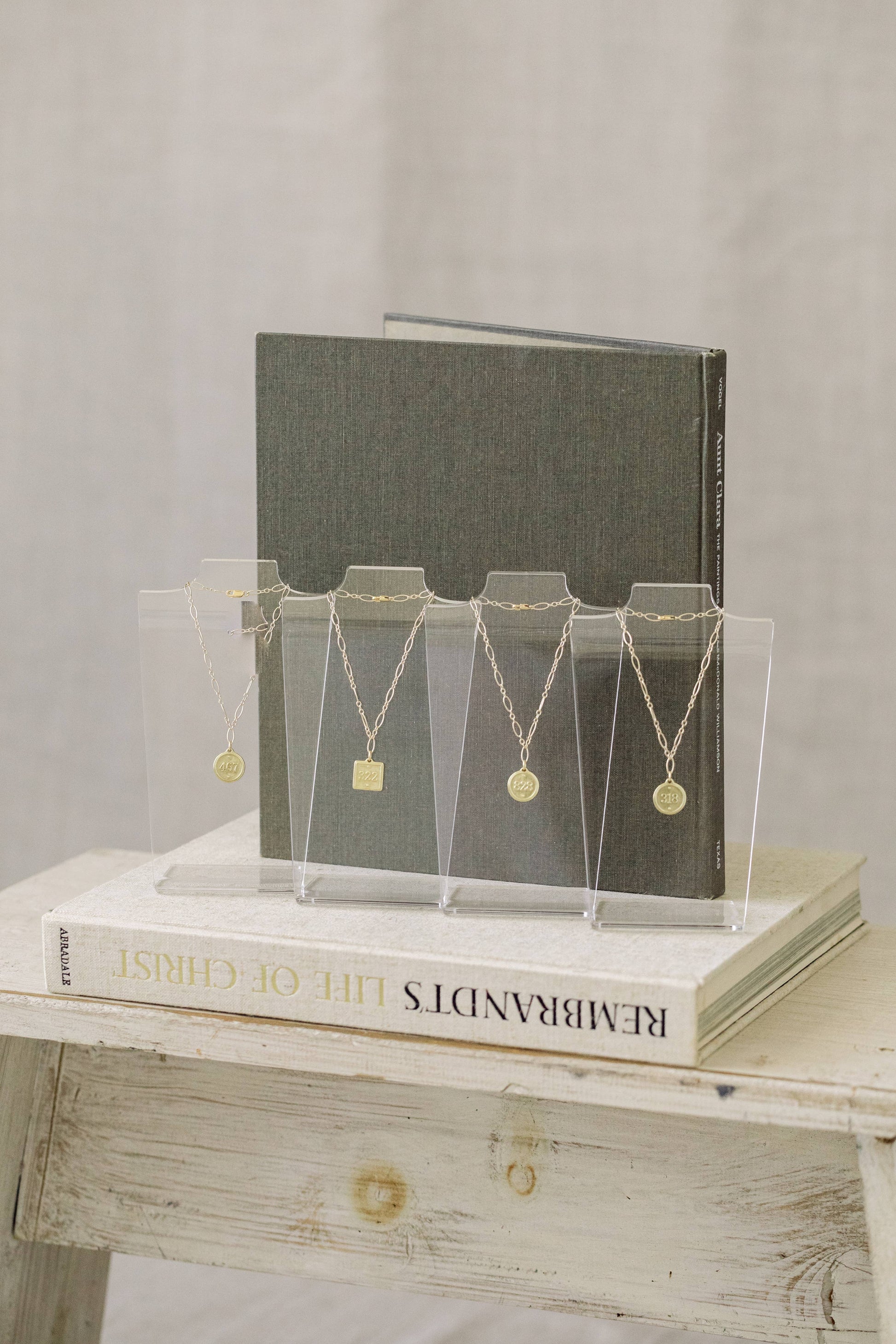 Five gold necklaces with round pendants displayed on clear acrylic stands, arranged on a white book in front of a dark gray book, set on a rustic white wooden table.