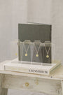 Five gold necklaces with round pendants displayed on clear acrylic stands, arranged on a white book in front of a dark gray book, set on a rustic white wooden table.