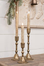 Three rustic gold-toned metal candlestick holders with cream taper candles, styled on a wooden surface against a white shiplap wall with holiday decor.