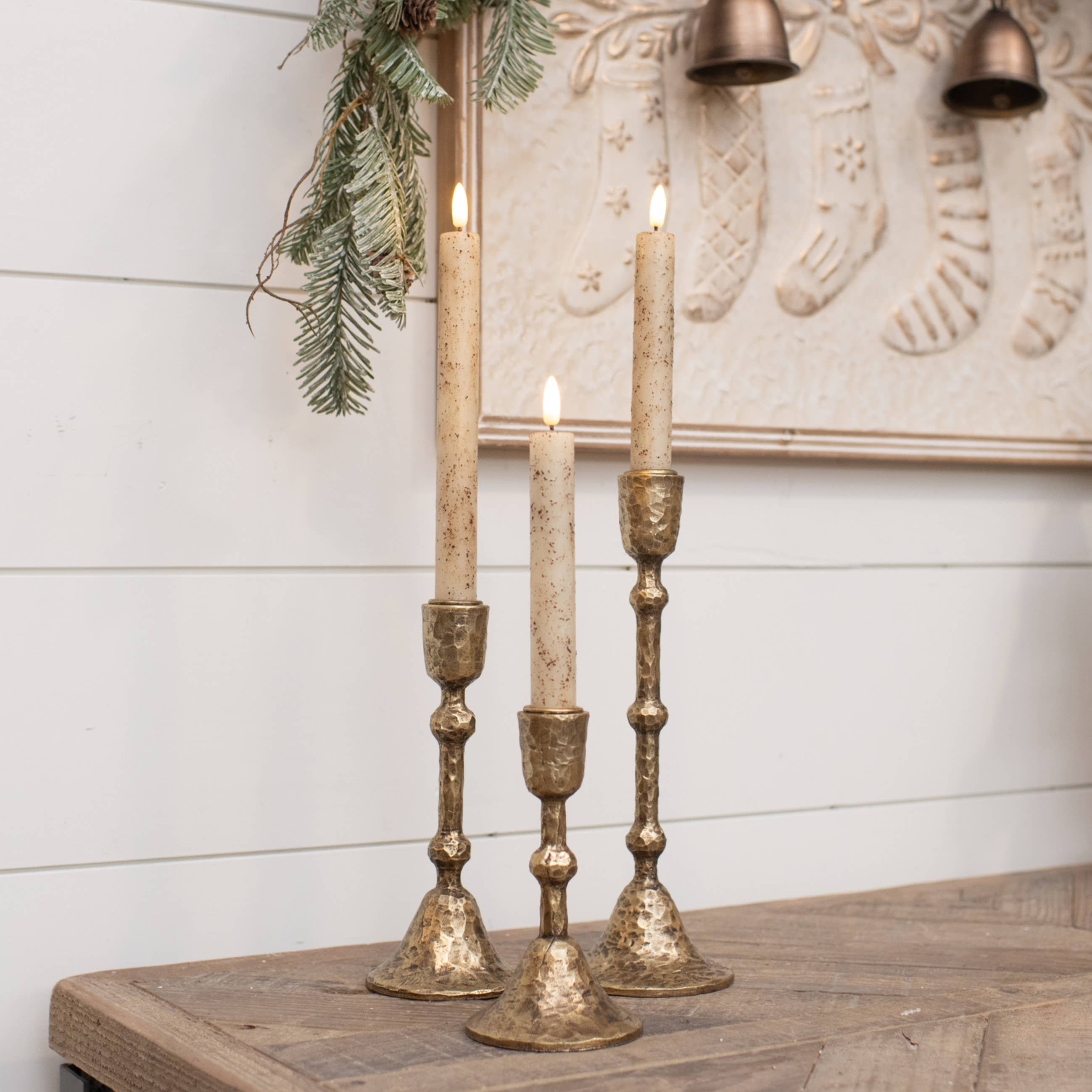 Three rustic gold-toned metal candlestick holders with cream taper candles, styled on a wooden surface against a white shiplap wall with holiday decor.