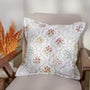 A square throw pillow with a white background and a colorful geometric pattern, resting on a beige upholstered chair.