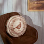 A round woven cushion with a cream and brown patterned design, resting on a brown leather chair against a white paneled wall.