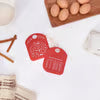Red shaped bowl scraper with Christmas design and measurements , arranged on a white surface with a wooden tray of eggs and a cinnamon stick in the background.