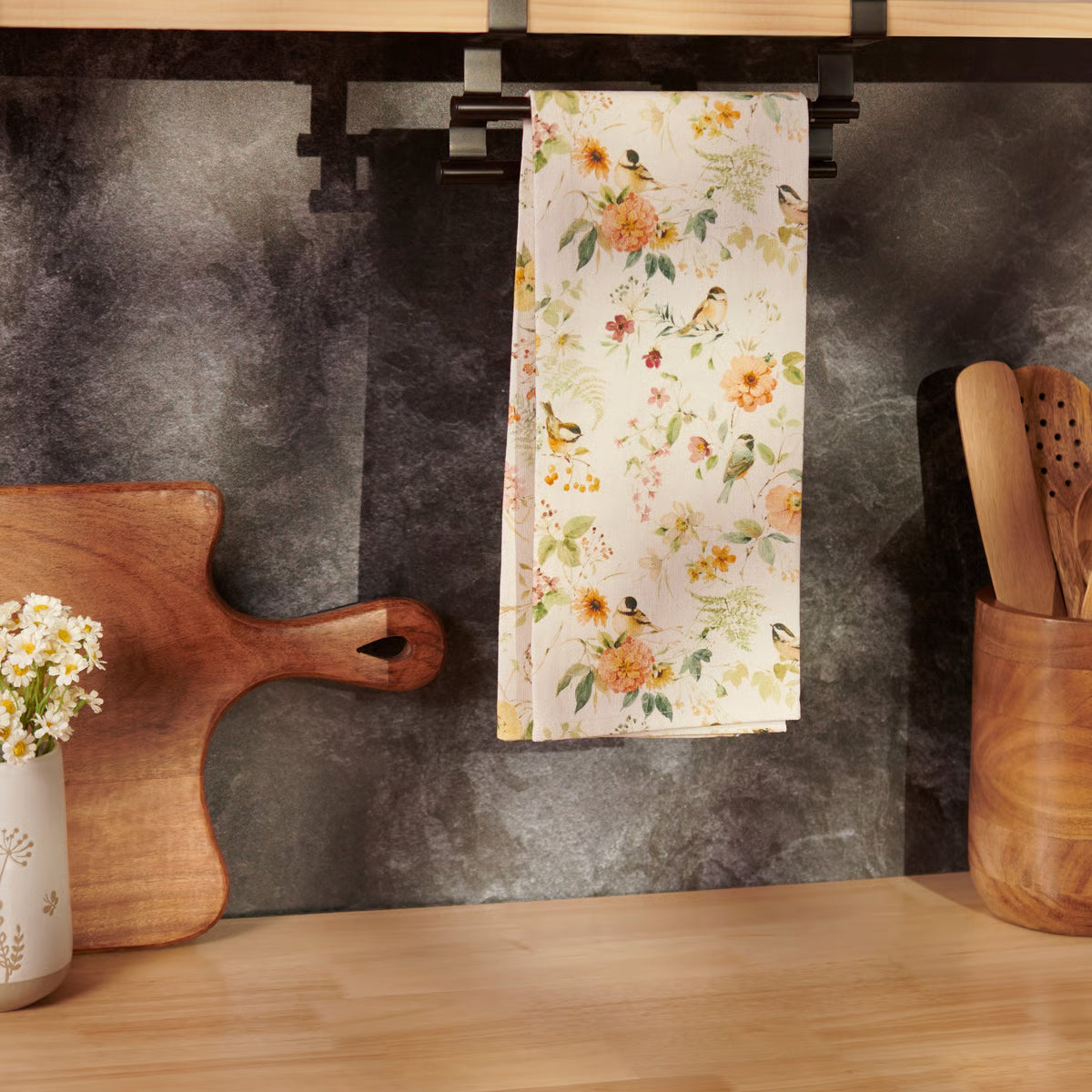 Floral-patterned towel hanging on a kitchen rack with wooden cutting board and utensils.