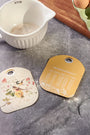 Two decorative kitchen labels on a gray surface with a measuring cup and eggs in the background.