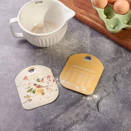 Two decorative kitchen labels on a gray surface with a measuring cup and eggs in the background.
