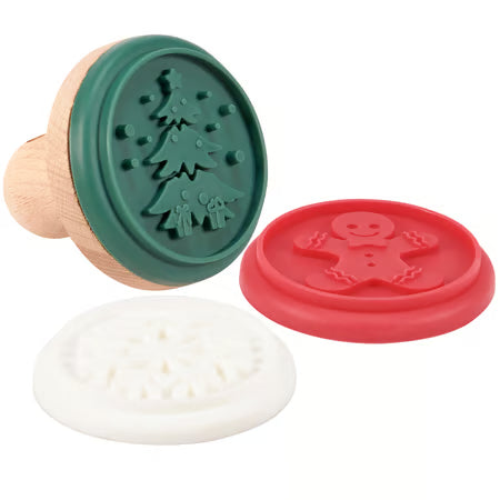 Three circular stamp-like molds or stamps shown on a plain white background: a wooden-handled green Christmas tree stamp, a red gingerbread man stamp, and a white snowflake stamp.
