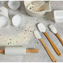 A set of silicone baking utensils with light brown wooden handles, including spatulas, a brush, a rolling pin, and cupcake liners, arranged on a gray stone surface with flour, sprinkles, and a mixing bowl in the background.