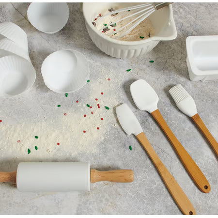 A set of silicone baking utensils with light brown wooden handles, including spatulas, a brush, a rolling pin, and cupcake liners, arranged on a gray stone surface with flour, sprinkles, and a mixing bowl in the background.