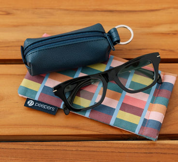 Black glasses on a colorful cloth with a blue case and Peepers label on a wooden surface
