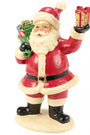 A painted Santa Claus figurine standing on a round base, wearing a red suit with white trim and holding a green wreath in one hand and a wrapped gift in the other, against a plain white background.