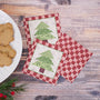 Three square fabric coasters with a Christmas tree and gingham pattern, arranged on a rustic wooden tabletop next to a plate of gingerbread cookies and evergreen sprigs.