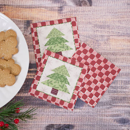 Three square fabric coasters with a Christmas tree and gingham pattern, arranged on a rustic wooden tabletop next to a plate of gingerbread cookies and evergreen sprigs.