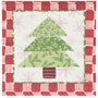 A square quilted napkin featuring a Christmas tree applique design. The tree is constructed from three layered green patterned triangles with pine branches and small white berries, set against a white background with light pink snowflakes.