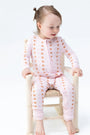A toddler girl sitting on a small wooden chair, wearing a light pink long-sleeve zip-up pajama set with a brown polka dot pattern. The photo is taken against a seamless white studio background.