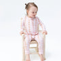 A toddler girl sitting on a small wooden chair, wearing a light pink long-sleeve zip-up pajama set with a brown polka dot pattern. The photo is taken against a seamless white studio background.