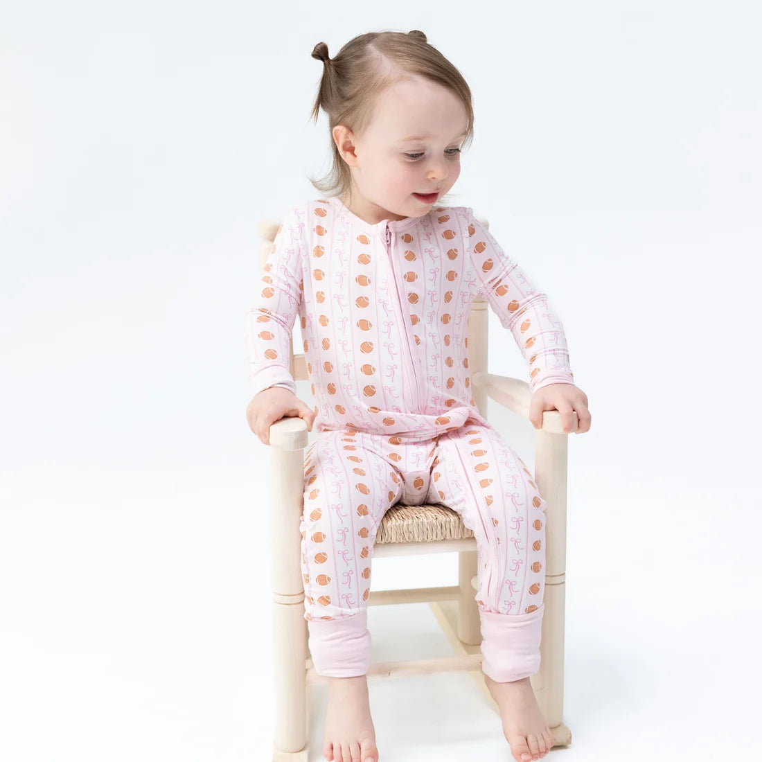 A toddler girl sitting on a small wooden chair, wearing a light pink long-sleeve zip-up pajama set with a brown polka dot pattern. The photo is taken against a seamless white studio background.