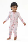A toddler standing barefoot against a plain white studio background, wearing a long-sleeve footless zip-up pajama or romper in light pink with a scattered floral print in soft red, blue, and green tones.