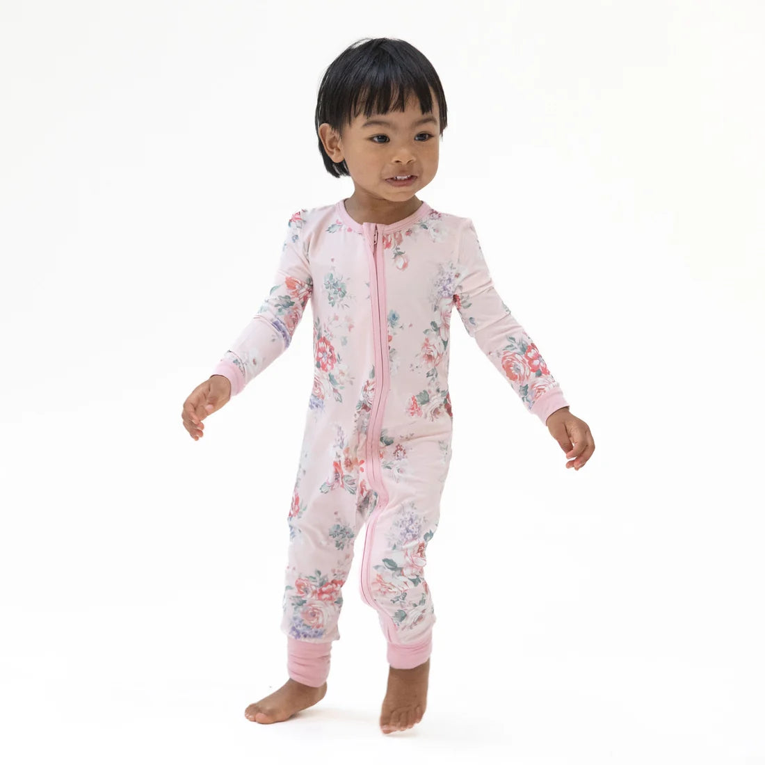 A toddler standing barefoot against a plain white studio background, wearing a long-sleeve footless zip-up pajama or romper in light pink with a scattered floral print in soft red, blue, and green tones.