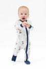 A baby standing on a white studio background wearing a white footless sleeper with navy blue trim and a teddy bear print, along with navy socks.