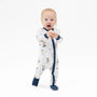 A baby standing on a white studio background wearing a white footless sleeper with navy blue trim and a teddy bear print, along with navy socks.