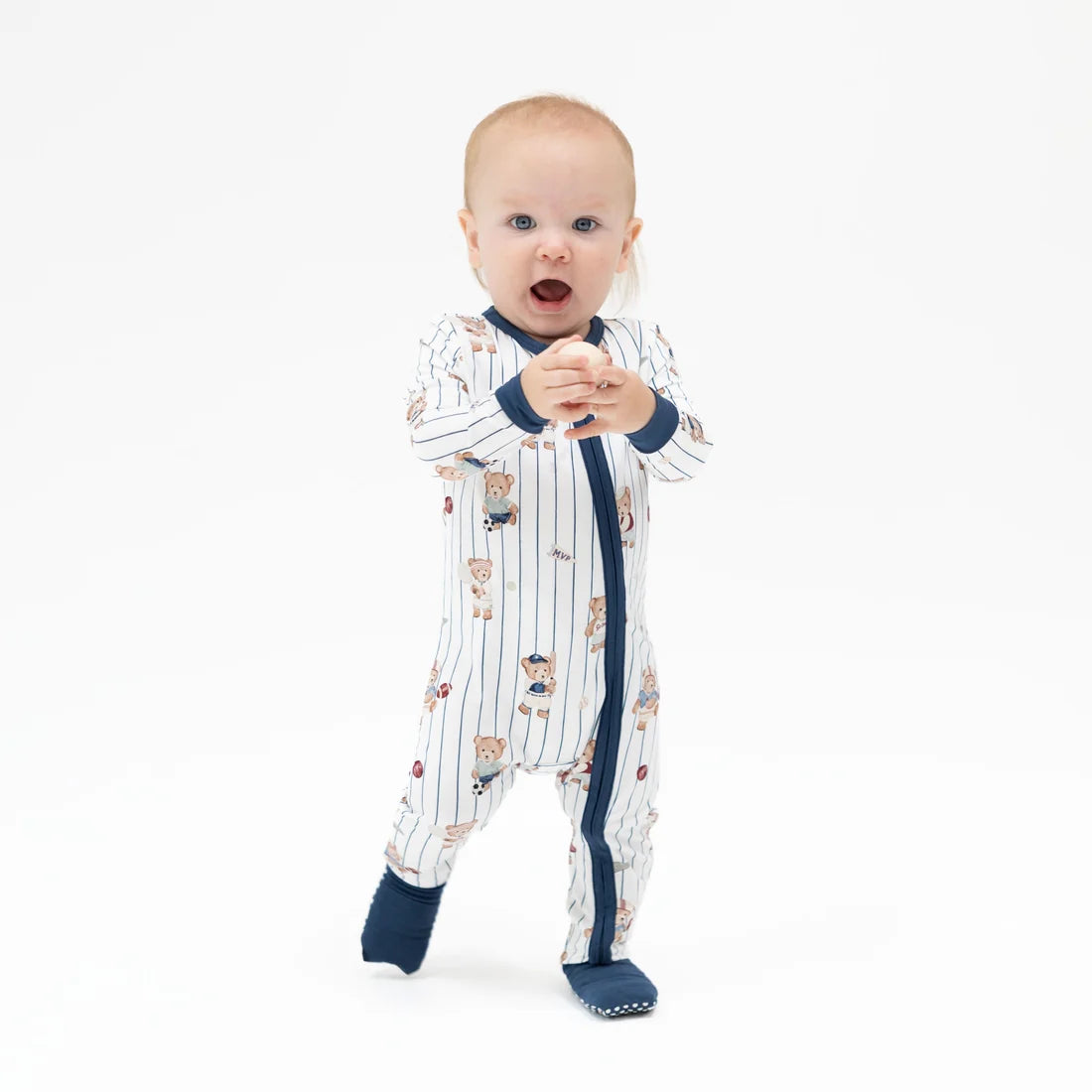 A baby standing on a white studio background wearing a white footless sleeper with navy blue trim and a teddy bear print, along with navy socks.
