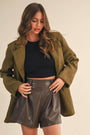 Woman wearing a green coat, black top, and leather shorts on a beige background