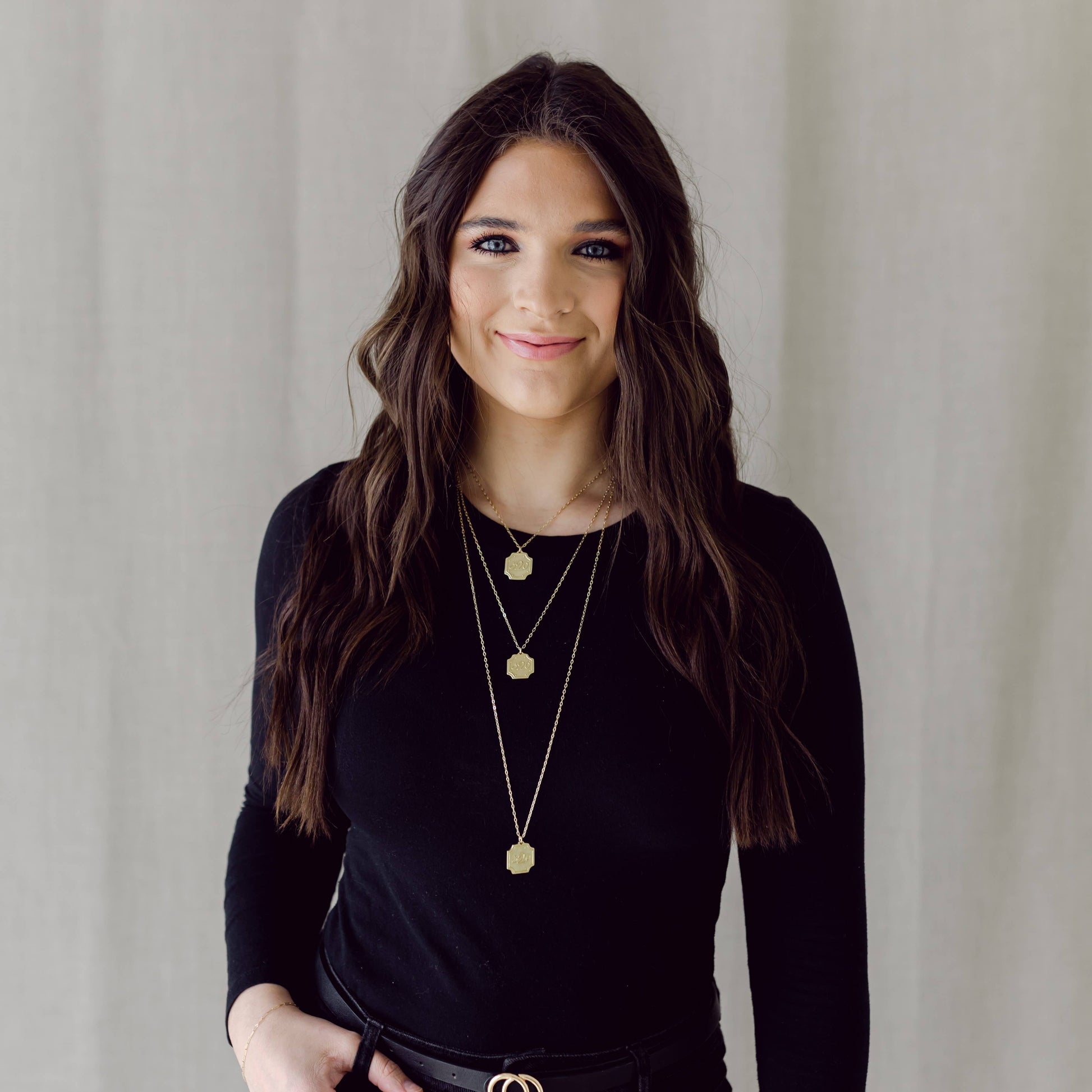 Woman wearing a black top and gold necklaces against a neutral background