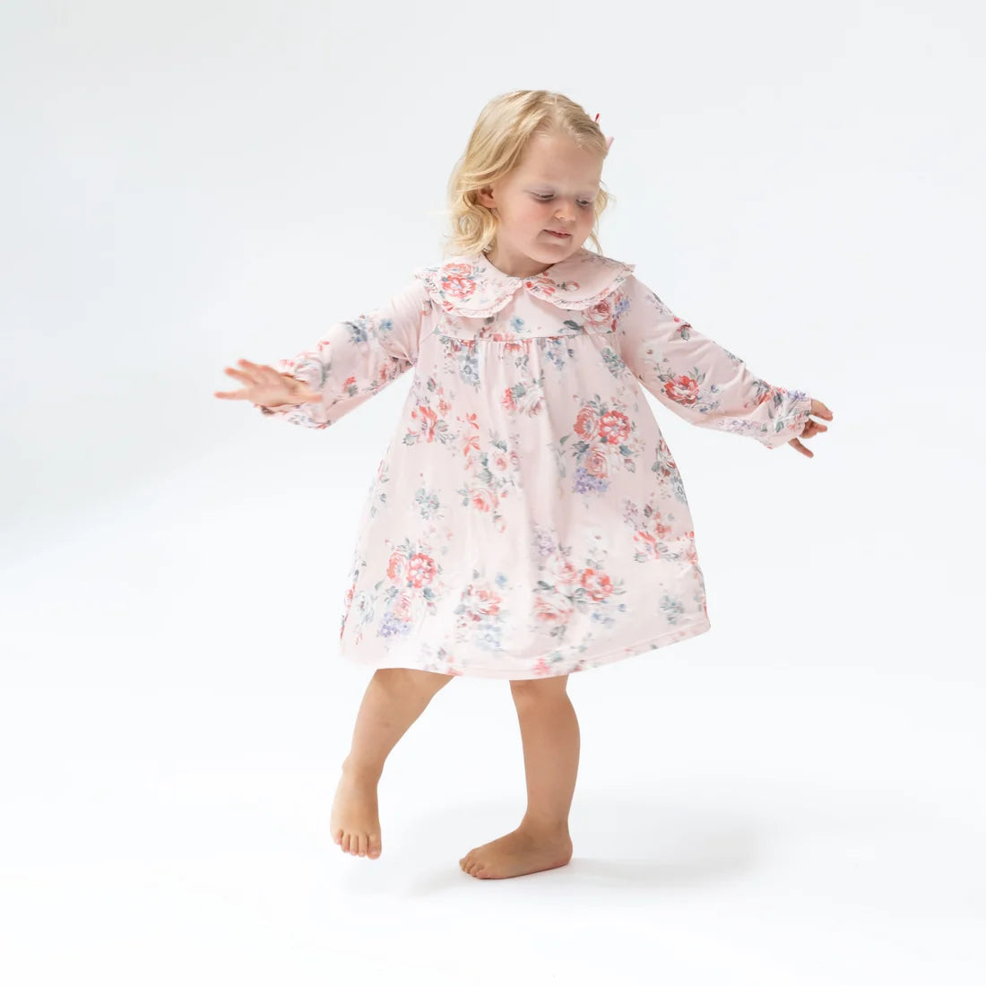 A young blonde girl wearing a light pink long-sleeve nightgown with a red and blue floral print, standing barefoot against a white studio background.
