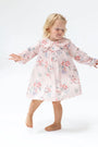 A young blonde girl wearing a light pink long-sleeve nightgown with a red and blue floral print, standing barefoot against a white studio background.