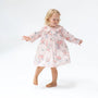 A young blonde girl wearing a light pink long-sleeve nightgown with a red and blue floral print, standing barefoot against a white studio background.