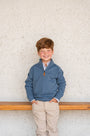 Young boy wearing a blue pullover with an orange zipper against a textured white wall.