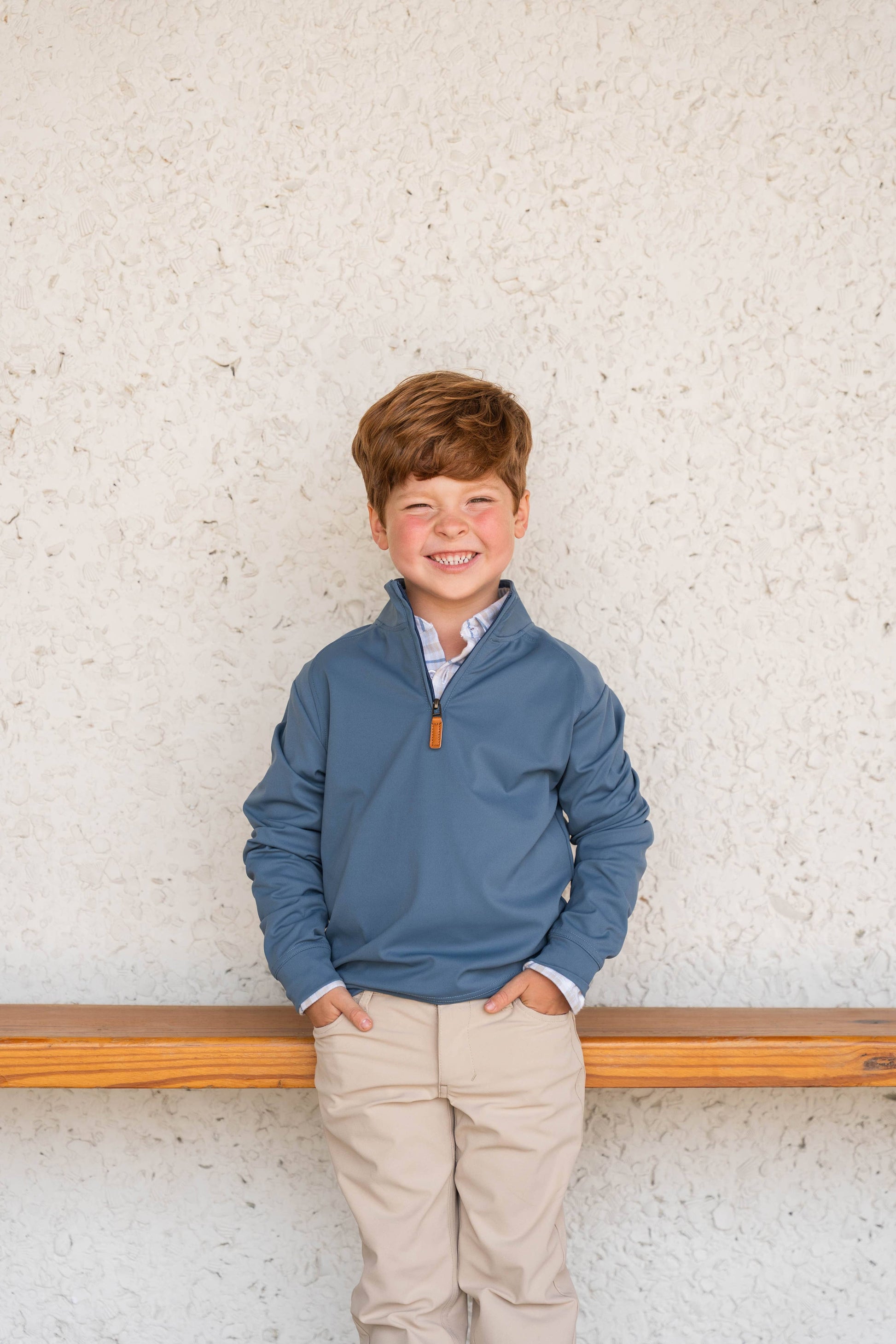 Young boy wearing a blue pullover with an orange zipper against a textured white wall.